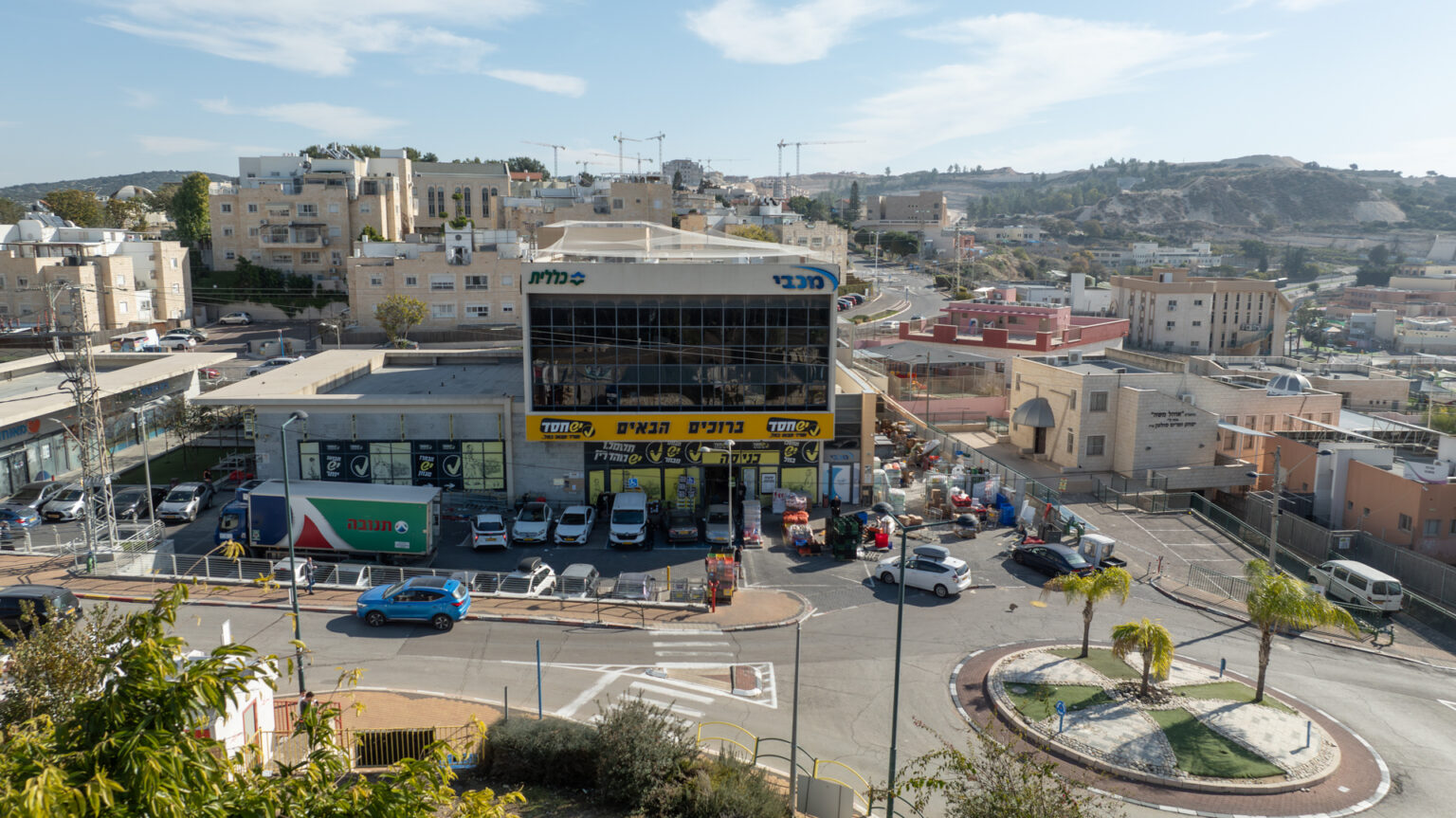 צילום חוץ של מבנה עם שלטים ״מכבי״,״כללית״, ו״ברוכים הבאים״ בניינים, כבישים, כיכר ומכוניות